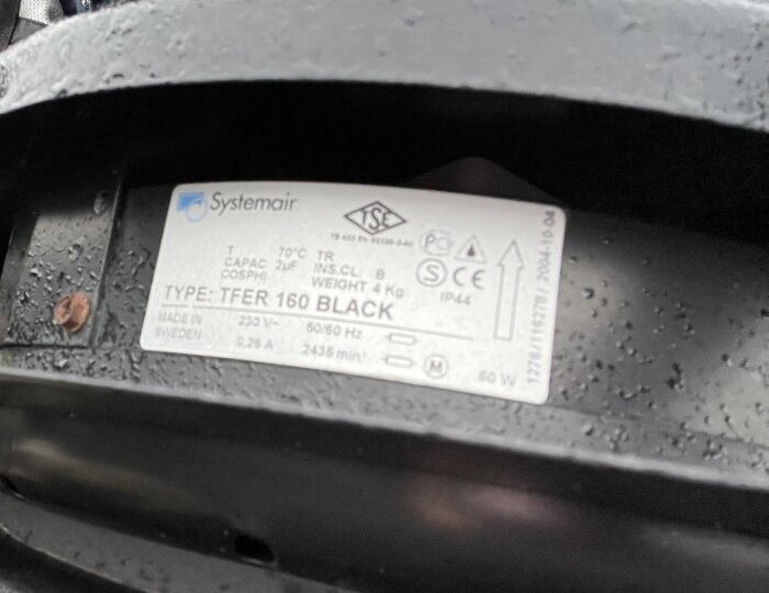 Närbild av en Systemair-produktetikett på en TFER 160 BLACK med tekniska specifikationer och märkningar, delvis täckt av vattenstänk.