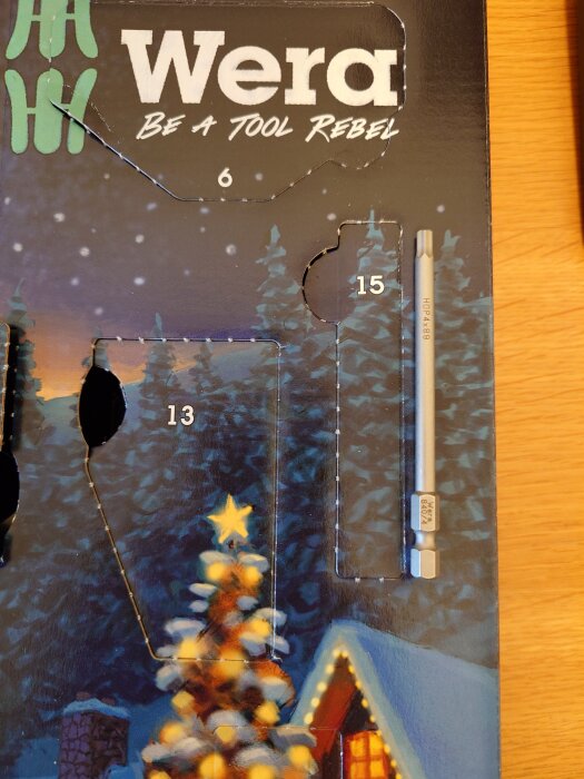 Wera kalender med öppnad lucka 15, avslöjar en verktygsbit. Bakgrund av julmotiv med snö och stjärna.