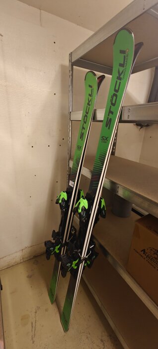 Gröna skidor av märket Stöckli, modell SX Laser, står lutade mot en vägg i ett förrådsutrymme.