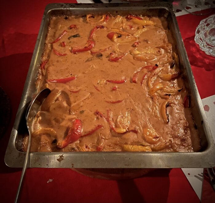 Krämig fläskfilégryta med paprika och svamp i en ugnsform, serverad på en röd duk, med en slev placerad till vänster.
