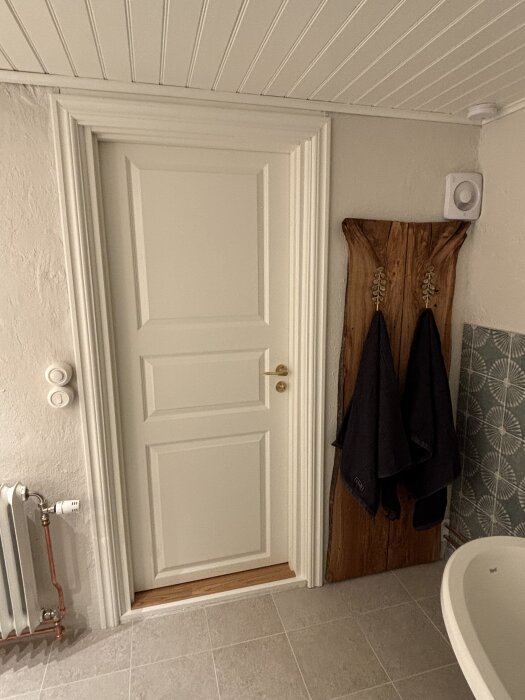 En vit dörr, en varmvattenradiator och ekskivor med svarta handdukar i ett delvis renoverat badrum med kakelvägg och ljusgrått klinkersgolv.