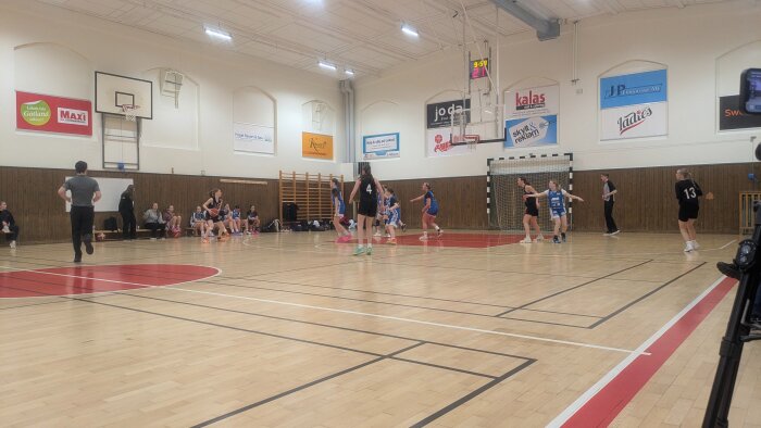Inomhus basketmatch med unga spelare i svart och blåa kläder på planen, tränare och åskådare vid sidan i sporthall.