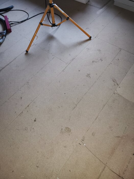 Golv under renovering med spånskivor täckta av fotspår och ett gult byggstativ i metall.