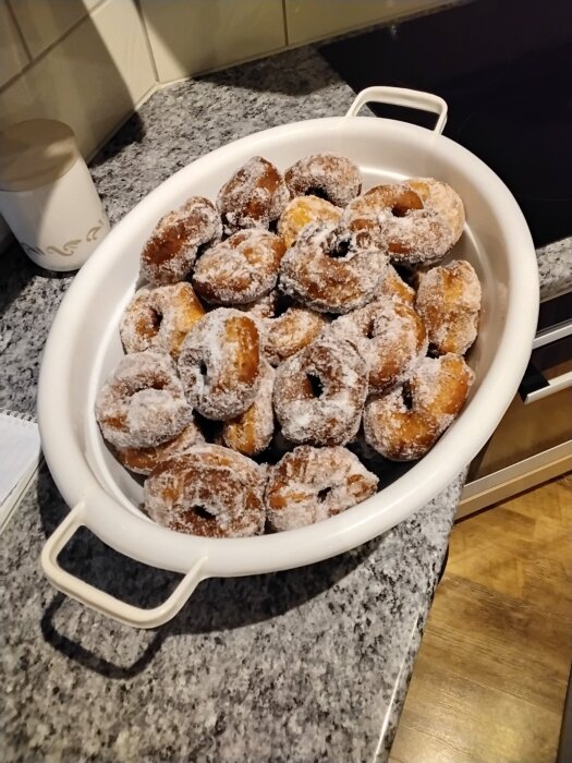 En vit skål fylld med nybakade, sockrade munkar står på en grå köksbänk.