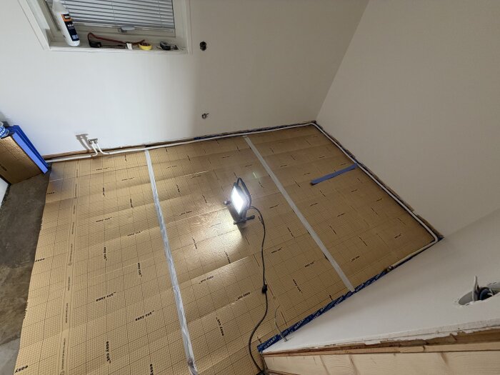 Rum under renovering med golvvärmeinstallation, strålkastare lyser på isoleringsmattor placerade på golvet, verktyg syns på fönsterbrädan.