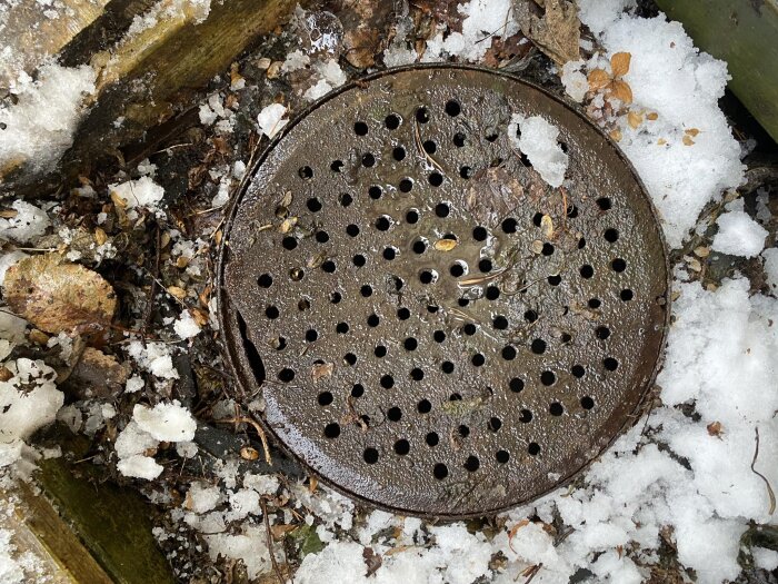 Brunnslock i metall med hål, omgiven av snö och löv, utanför ett radhus.