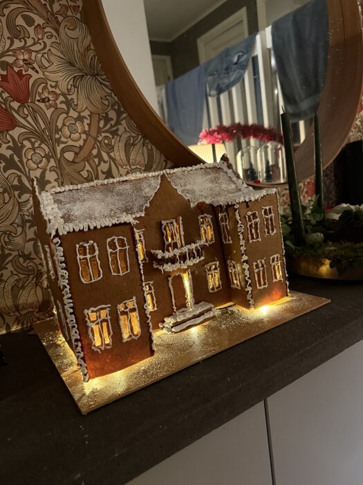 Pepparkakshus med snöliknande glasyr och ljus inuti, placerat på en hylla framför en spegel och tapet med blommönster.