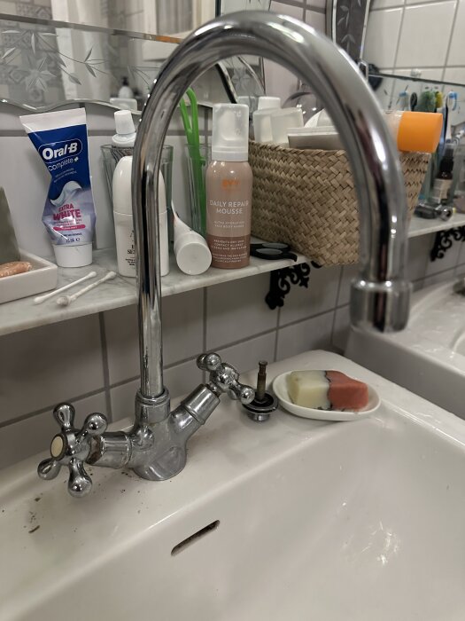 Kromfärgad köksblandare med två vred vid vask, en ventiltopp saknas. Bakgrunden har tandkräm och andra hygienprodukter på en hylla.