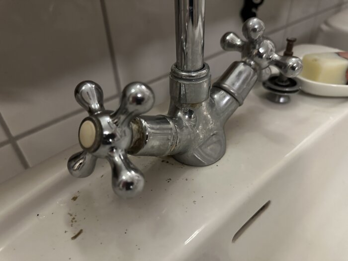 Kromad tvågreppsblandare med vita detaljer, installerad i ett handfat. Ingen märkning synlig på blandaren. Eventuellt behov av reservdelar.
