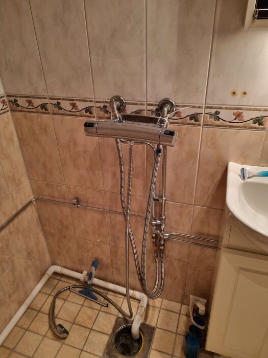 Badrum med kakelvägg, duschblandare och exponerad golvbrunn. Fråga om installation av duschkabin över brunnen.