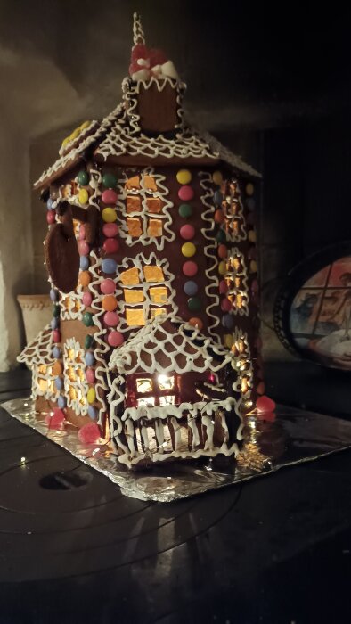 Dekorerat pepparkakshus med glasyr, färggranna godisar och kristyrdetaljer, med små lampor inuti som lyser upp fönstren.