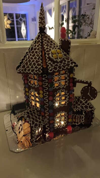 Pepparkakshus med dekorationer av glasyr och godis, upplyst inifrån, placerad på en folieklädd bricka framför ett fönster med julpynt.