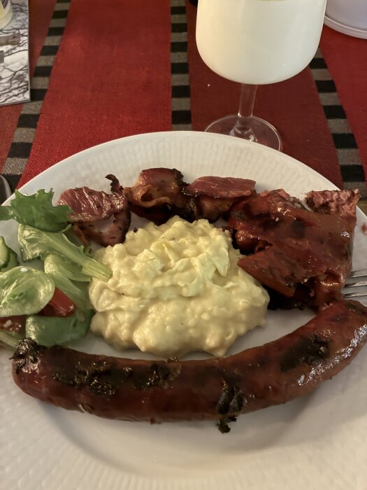 Tallrik med grillat kött, korv, potatissallad och sallad. Glas med mjölk i bakgrunden på en röd duk.