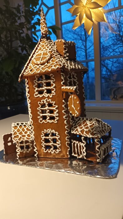 Mumininspirerat pepparkakshus med vitt kristyrmönster och klockdetalj, placerat på folie i ett hem med en juldekorationsstjärna i bakgrunden.