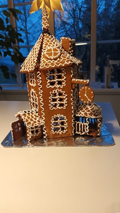 Mumininspirerat pepparkakshus med vit glasyr, detaljerad utsmyckning och stjärna på toppen, mot fönsterbakgrund.