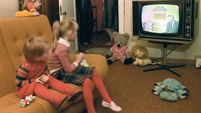 Barn tittar på TV i ett vardagsrum, äldre modell på TV:n och leksaker på golvet.