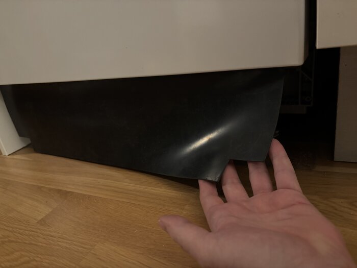 En hand lyfter en svart gummimatta med vaddering som hänger ner bakom en apparatlucka, undrar om den ska tas bort.