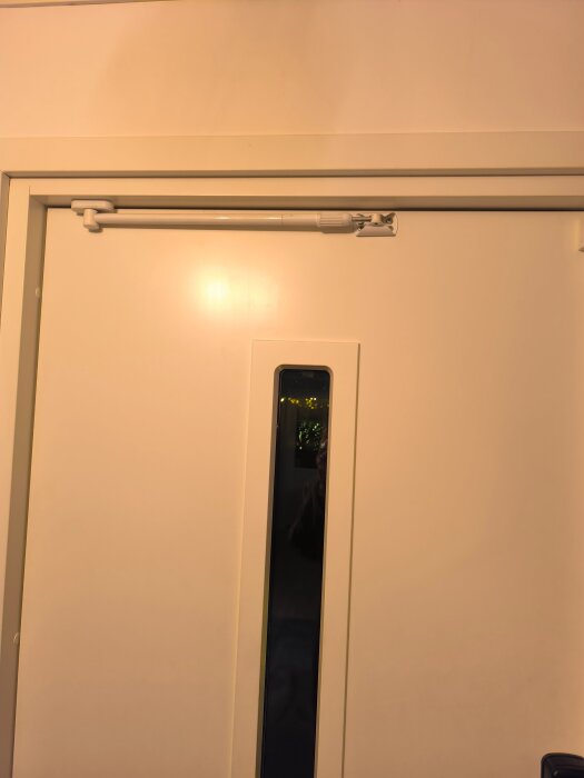 Vit dörr med en dörrbroms monterad upptill. Dörrbromsen ser ut att vara en modell som förhindrar dörren från att blåsa upp, ej slå igen.