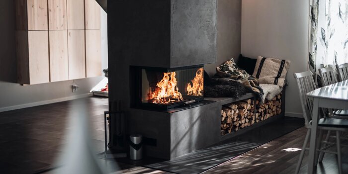 Inbyggd kamin med eld och glasfront, intill en bänk med plädar och vedförvaring under, i ett modernt rum med trädetaljer.