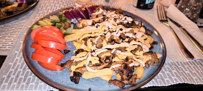 Kebabtallrik med pommes, kött, vit och röd sås, serverad med skivad tomat, gurka och rödlök på en blå tallrik.