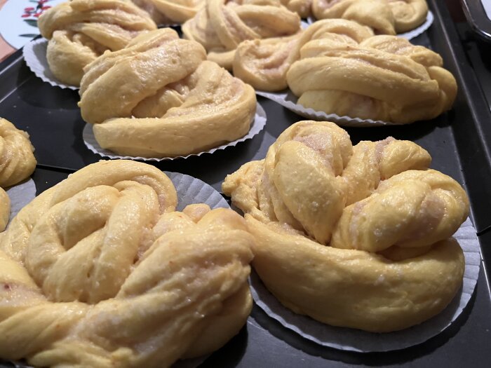 Lussebullar med smör- och mandelfyllning på en bakplåt. Bullarna har en gyllengul färg och ligger i papperformar.