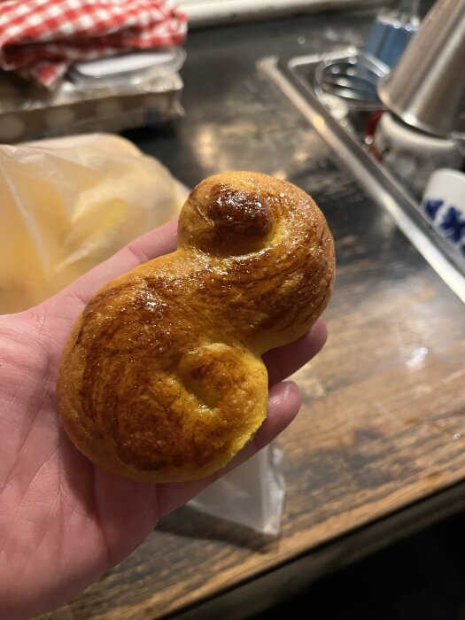 Hand håller en lussekatt med glansig yta, bakad med smör och mandelfyllning, mot en bakgrund av kökshandduk och ugnsplåt.
