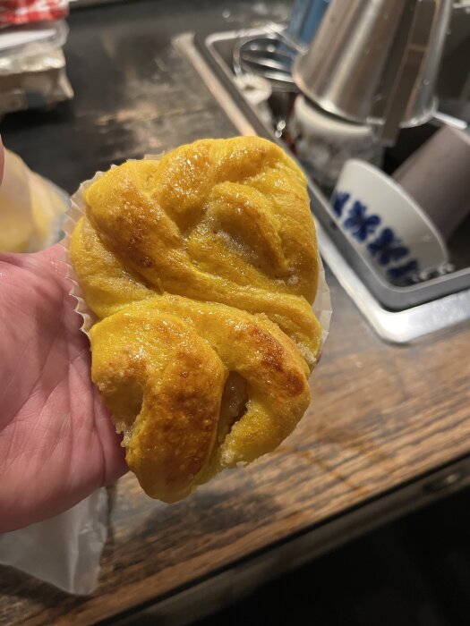 Lussekatt med smör- och mandelfyllning, något tillplattad men gyllene och glansig, ligger i en hand ovanför en köksbänk.