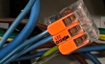 Elektriska ledningar anslutna till en Wago-kopplingsklämma, med orange vipparmar, i ett elskåp.