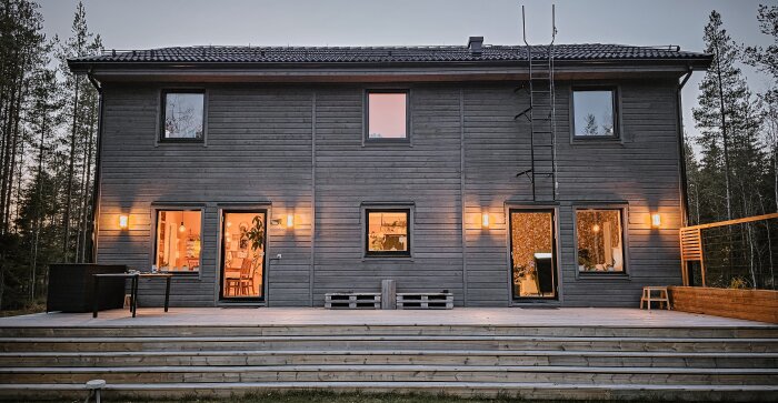 Tvåvåningshus med fasadbelysning i skymning, stora fönster och bred trappa med träaltan, omgiven av tallar.