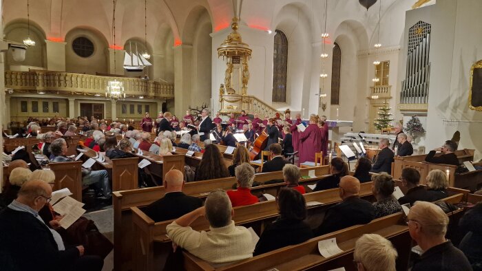 Kyrkokonsert med kör i lila kläder som sjunger framför en fullsatt publik i en vackert dekorerad kyrka med orgel och julgran.