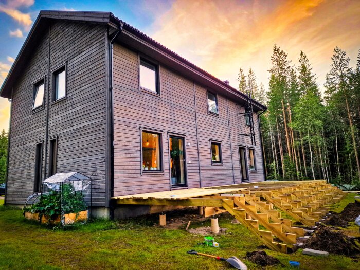 Trähus med nybyggd trätrappa och altan under konstruktion, intill en skog vid solnedgång.