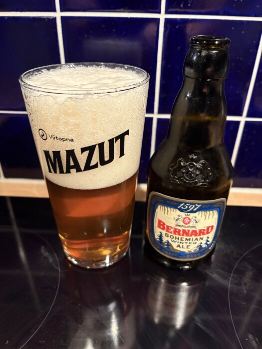 En glas med skummande öl bredvid en flaska Bernard Bohemian Winter Ale, 8,2% alkoholhalt, mot en blå kakelbakgrund.