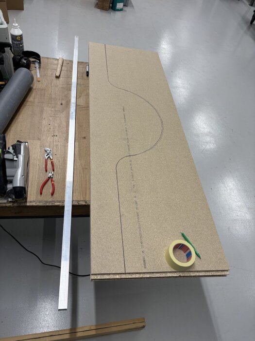 MDF-skiva med utritad form för hjulhus, omgiven av verktyg och material, på ett arbetsbord i verkstad.