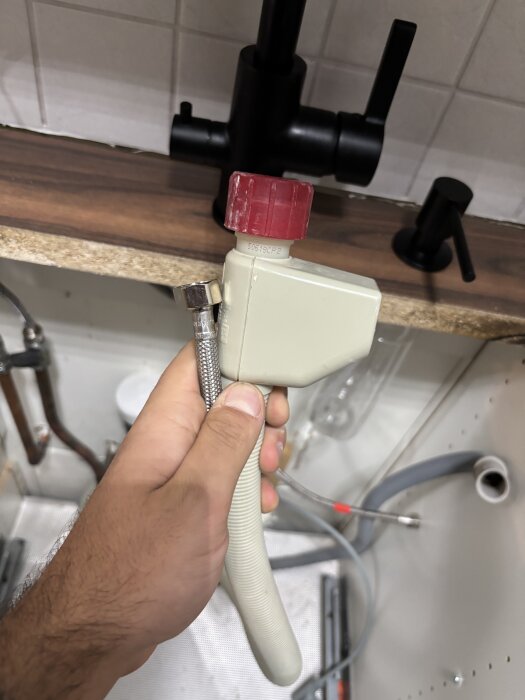 Hand håller i en koppling för köksblandare med en röd ventil i en diskbänksinstallation.