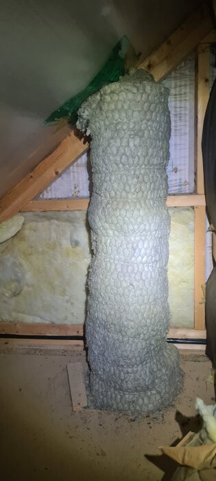 Isolerat ventilationsrör i träkattvind under snedtak. Rör böjt och har ojämn ytstruktur, kringliggande träregel och isoleringsmaterial synliga.