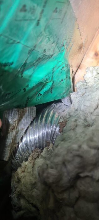 Ventilationsrör av aluminium i trångt utrymme, omgivet av grönt material och isolering. Bucklig yta på röret syns tydligt.