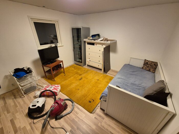 Rum med säng, byrå, fönster, TV på ett bord, klädförvaring och dammsugare på golvet. Ljus parkett och gul matta.