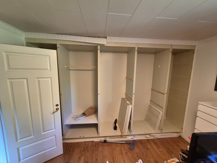 Öppen garderob under montering med vita dörrar och hyllor, verktyg och material ligger på golvet i ett rum med parkettgolv.