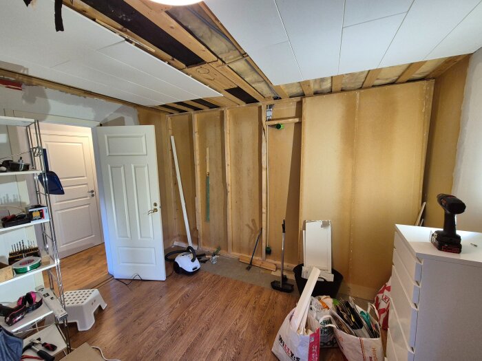 Rum under renovering med synliga träreglar, vita dörrar, verktyg och material spridda på golvet och hyllor i bakgrunden.
