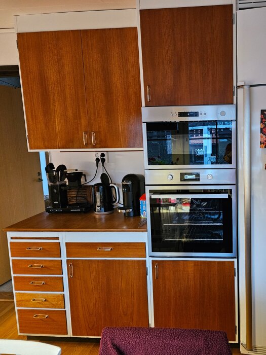 Inbyggd ugn och mikrovågsugn i ett gammalt köksskåp med träluckor och lådor.