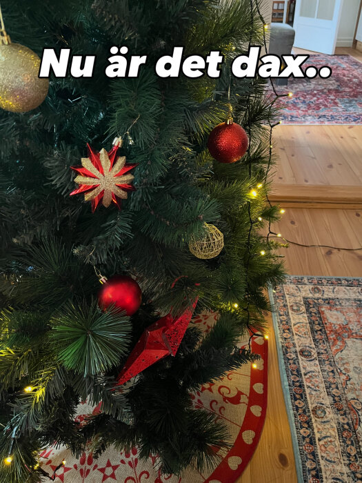 Julgran dekorerad med röda och gyllene kulor, ljusslingor och stjärnor, i ett rum med trägolv och orientaliska mattor. Texten säger "Nu är det dax..".