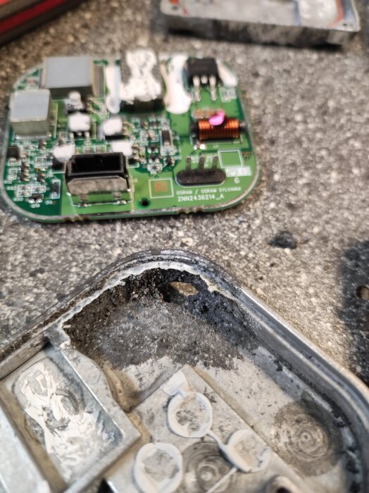 Grön kretskort med elektroniska komponenter jämte en del av ett slitet metallhölje på en grå yta.