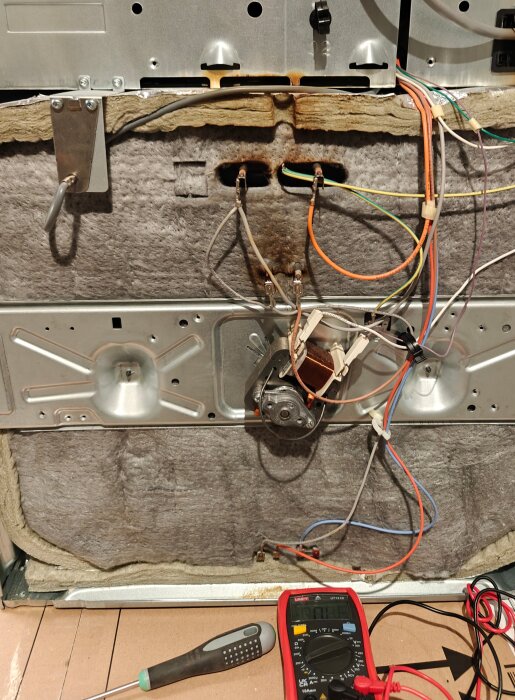 En trasig mikrovågsugn, med baksidan avtäckt, visar blottlagda ledningar och en motor; en multimeter ligger på golvet bredvid.