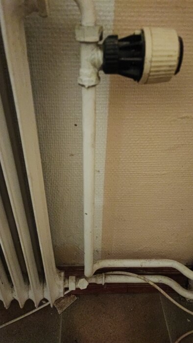 Radiatorrör med termostatventil mot beige vägg, anslutet till ett element och golv med klinkerplattor.