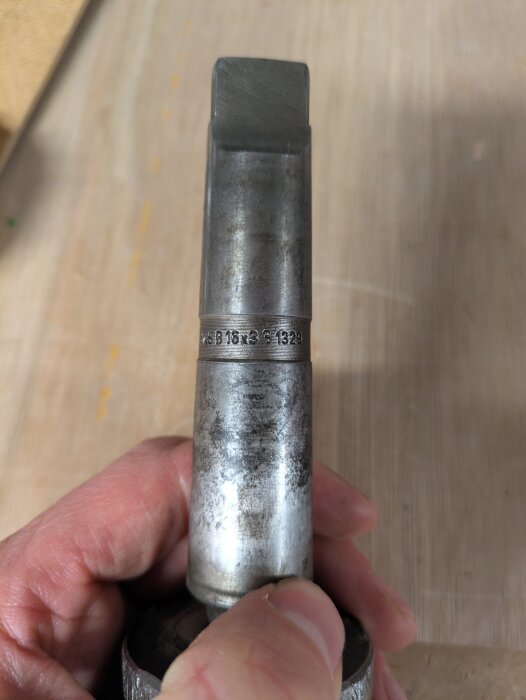 Hand som håller en metallcylinder med ingraverad text "MAS B 16x3 (krummelur) 1329". Verktyget diskuteras för ersättning.