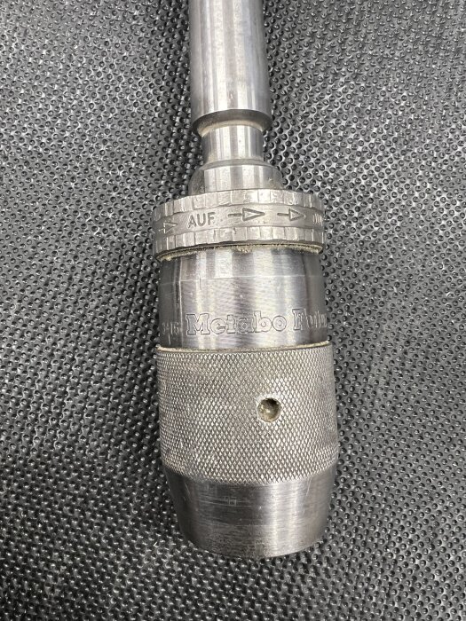 Metabo chuck med märkningar ”3-16” och ”1/8-5/8”, monterad på en axel, placerad på en svart perforerad yta.