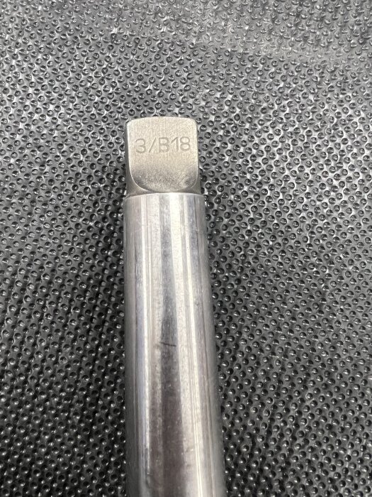 Axel av metall märkt "3/B18" på ett prickigt underlag.