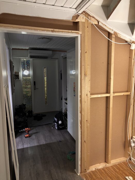 Öppen vägg i hallen på 70-tals kedjehus, med synliga reglar och plywood, trappa till höger. Synlig borrmaskin och kläder på golvet.