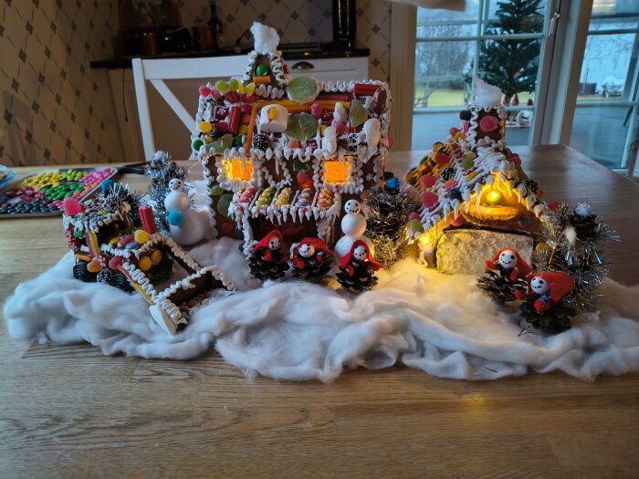 Pepparkakshus dekorerade med godis och glasyr, omgivet av små snögubbar och granar gjorda av kottar, står på ett bord med bomullssnö.