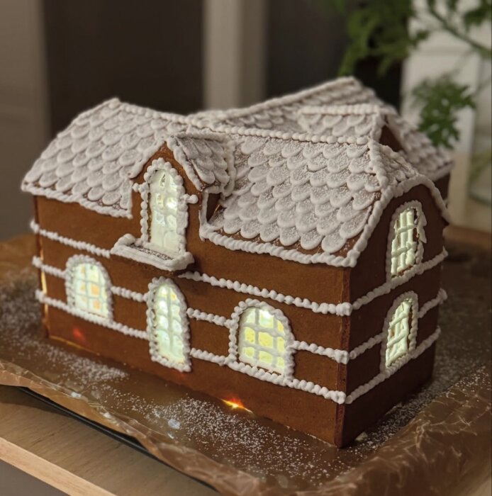 Dekorerat pepparkakshus med snötäckt tak, fönster med belysning och detaljerade icinglinjer, inspirerat av ett sekelskifteshus.